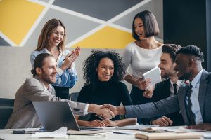 Two confident businessmen shaking hands while sitting with their colleagues at the office desk Two confident businessmen shaking hands while sitting with their colleagues at the office desk